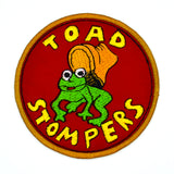 Circular patch on red fabric and gold border featuring a toad being stepped on by a boot and the words 'toad stompers' stitched in yellow thread, as a reproduction from the sci-fi movie Hell Comes to Frogtown.
