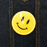 A circular yellow embroidered patch of a smiley face with a white splatter across its right eye.