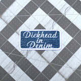 A rectangular embroidered patch on denim fabric with a white border and white lettering that reads, 'Dickhead in denim'.