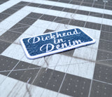 A rectangular embroidered patch on denim fabric with a white border and white lettering that reads, 'Dickhead in denim'.