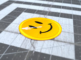 Close up detail of a circular yellow embroidered patch of a smiley face with a white splatter across its right eye.