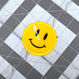 A circular yellow embroidered patch of a smiley face with a white splatter across its right eye.