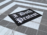Close up detail of a rectangular black embroidered patch with white Gothic lettering that reads 'I Doth Protest'.