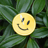 A circular yellow embroidered patch of a smiley face with a white splatter across its right eye.