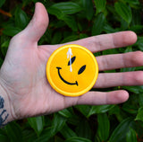 A circular yellow embroidered patch of a smiley face with a white splatter across its right eye. Shown in the palm of a hand for size reference.