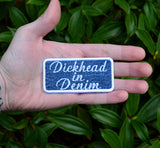 A rectangular embroidered patch on denim fabric with a white border and white lettering that reads, 'Dickhead in denim'. Held in the palm of a hand for size reference.