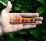 An embroidered rectangular patch with the text 'BIGGUS DICKUS' in cream lettering on a red background, bordered in gold. Held in the palm of a hand for size reference.