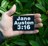 Rectangular black embroidered patch with white block lettering that says, 'Jane Austen 3:16". Held in the palm of a hand for size reference.