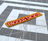 Close up detail of an embroidered rectangular patch with the text 'BIGGUS DICKUS' in cream lettering on a red background, bordered in gold.