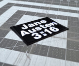 Close up detail of a rectangular black embroidered patch with white block lettering that says, 'Jane Austen 3:16".