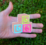 An embroidered patch resembling three children's building blocks showing the number six in three different colors. Shown in the palm of a hand for size reference.