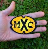 A black and yellow oval-shaped logo patch for the comedy tv show MXC. Held in the palm of a hand for size reference.