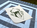 Close up detail of a rectangular off-white fabric patch featuring a dark green graphic print of an antlered figure holding a dagger, surrounded by a diamond border.
