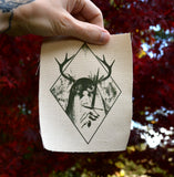 A rectangular off-white fabric patch featuring a dark green graphic print of an antlered figure holding a dagger, surrounded by a diamond border. Shown being held up by a hand for size reference.