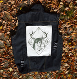 A rectangular off-white fabric patch featuring a dark green graphic print of an antlered figure holding a dagger, surrounded by a diamond border. Shown on the back of a denim vest.