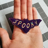 Purple bat-shaped felt patch with the word spooky sewn in multicolor thread, shown in a hand for size reference.