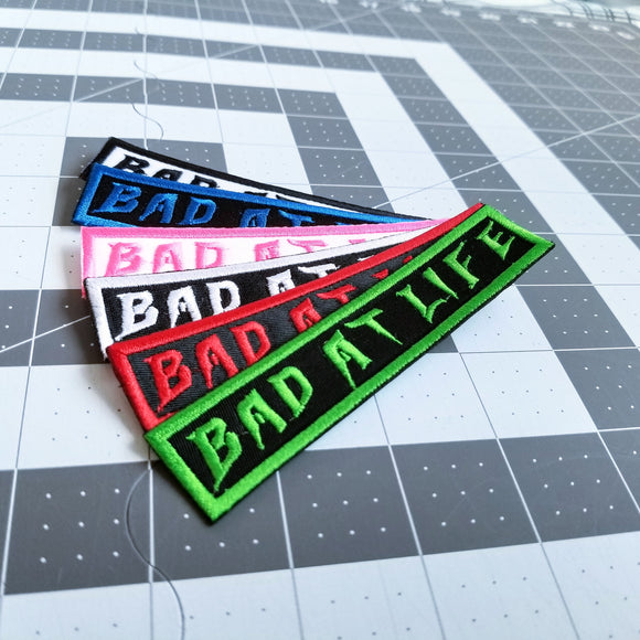 A collection of rectangular embroidered patches with the phrase bad at life in various colors.
