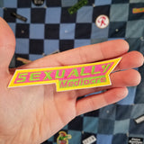 A sharp angled rectangular embroidered patch in neon green, pink, and yellow that says, 'Sexually mediocre'.