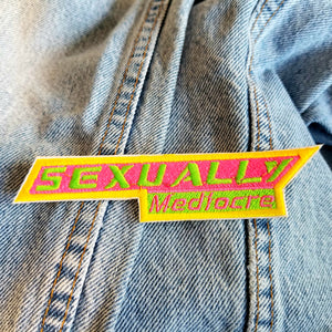 A sharp angled rectangular embroidered patch in neon green, pink, and yellow that says, 'Sexually mediocre'.