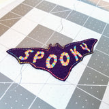Purple bat-shaped felt patch with the word spooky sewn in multicolor thread.