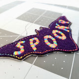 Close-up detail of a purple bat-shaped felt patch with the word spooky sewn in multicolor thread.