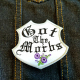 A white emblem-shaped and silver bordered embroidered patch with purple flowers and the words Got the Morbs in old english lettering.