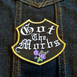 A black emblem-shaped and gold bordered embroidered patch with purple flowers and the words Got the Morbs in old english lettering.